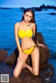 A woman in a yellow bikini sitting on a rock by the water.